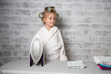 a little girl in curlers strokes, stands at the ironing board and smiles. Gray brick background,...