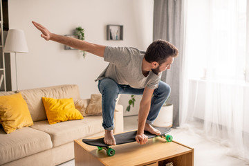 Carefree young bearded man skateboarding in crouched pose on coffee table in living room during home isolation © pressmaster
