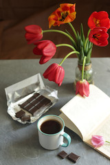 On the table is a cup of coffee, chocolate bar, a book and flowers.

