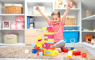 Smiling little girl sitting on the floor in the kids room and holding her hands up while playing with toy blocks