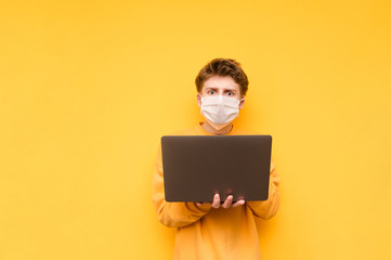 Surprised young man in a medical mask stands with a laptop in his hands and looks at the camera with a shocked face on a yellow background. Coronavirus pandemic. COVID-19. Quarantine.