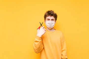 Portrait of a funny shaggy young man with scissors in his hands and a medical mask on his face stands on a yellow background and looks at the camera with surprised eyes. Quarantined haircut.