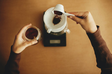 Hands pouring grounded coffee in filter pour over on glass kettle on scale,top view. Preparing for alternative coffee brewing v60. Person holding spoon with grind coffee on brown background