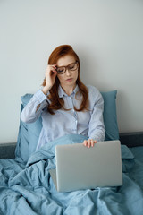 A red-haired girl in her pajamas sits in bed and closes her laptop, taking off her glasses.