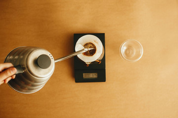 Alternative coffee brewing v60. Barista making filter coffee. Hands pouring hot water from steel kettle in filter with ground coffee in pour over on scale on brown background.