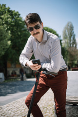 Handsome man with sunglasses and moustache leaning on scooter. Young man with facial hair looking at camera while holding phone and propping himself up on scooter.
