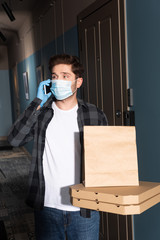 Fototapeta na wymiar Delivery man in medical mask holding pizza boxes and package while talking on smartphone on entryway