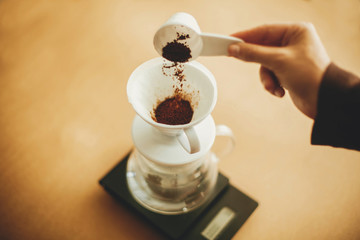 Hands pouring grounded coffee in filter. Preparing for alternative coffee brewing v60. Person holding spoon with grind coffee on background of glass kettle with pour over on scale