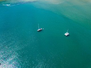 A view from a high-angle drone sees sailboats leaving anchors in a beautiful bay, clear glass-like water.