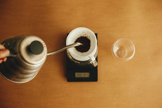 Pouring Hot Water From Steel Kettle In Filter With Ground Coffee In Pour Over On Scale, Top View. Alternative Coffee Brewing V60. Barista Making Filter Coffee On Brown Background