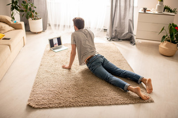 Rear view of man in jeans practicing cobra pose while watching online yoga class on laptop at home