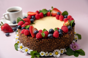 Cheesecake decorated with berries and flowers on a light pink background with a cup of coffee on the left side.