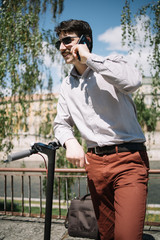 Portrait of guy in elegant clothes speaking on the phone. Happy boy with moustaches wearing sunglassesand talking on mobile while leaning on electric scooter.