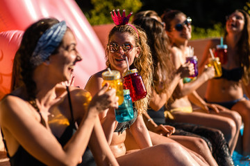 Group of girlfriends at a poolside summer party sitting at the edge of a swimming pool drinking and having fun.