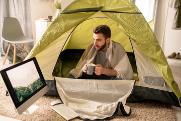 Serious young man under blanket sitting with mug in camping tent and looking at forest picture on...