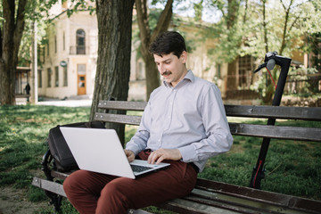 Businessman sitting on bench in park and using laptop. Male freelancer in elegant outfit working on computer while sitting on wooden bench in city park.