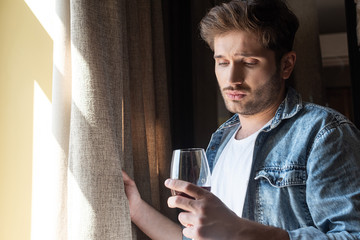 Upset man holding glass of wine near window in living room