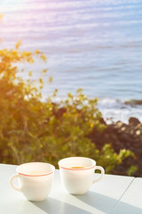 Two cups of coffee on a balcony with a view to the ocean and trees on the shore. Sunny beautiful morning. Background in soft focus.