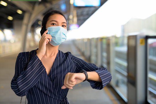 Young Asian businesswoman with mask thinking while talking on the phone and checking smartwatch at the station