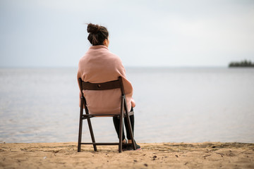 
a lonely girl sits on a wooden chair on a sandy beach by the lake and looks into the distance, a...
