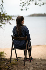 Fototapeta premium a young girl sits on a chair near a pond, against the background of a lake or river or sea, her back to the camera, looks into the distance, admires the view and enjoys the fresh air, calm and silence