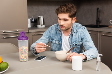 Handsome man holding thermometer near smartphone and cereals on kitchen table