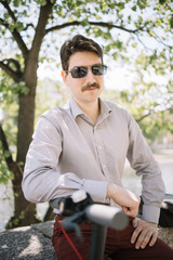 Handsome man with moustache and sunglasses posing under tree. Portrait of man with moustache wearing shirt and sunglasses while leaning on bike rudder.