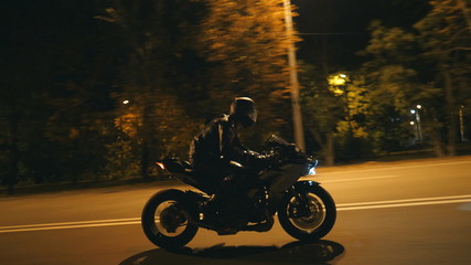 Young man in helmet riding fast on modern sport motorbike at evening city street. Motorcyclist racing his motorcycle on night empty road. Guy driving bike. Concept of freedom and hobby. Side view