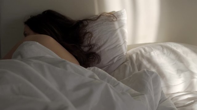Woman sleeping in bed in the morning, dark brown long hair on pillow, sunlight falling on bed.