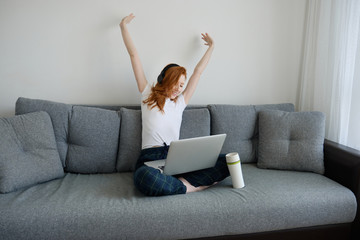 Fototapeta na wymiar A red-haired girl sits on a sofa in home clothes with a laptop on crossed legs and stretches her arms to the sides