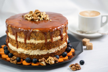 Cup of coffee with two pieces of sugar on a saucer and next to a carrot cake covered with caramel and decorated with nuts on top, behind a white textile background