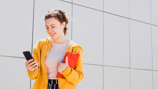 Young happy millennial woman in headphones using mobile phone for video call. Smiling student teenager watching video, webinar with smartphone,using wireless Internet connection outdoor