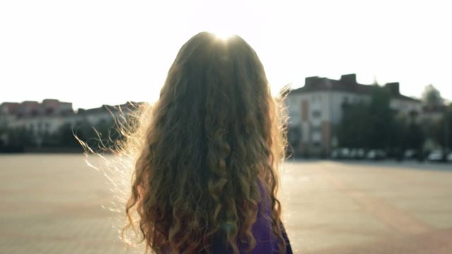 Curly long haired girl without makeup turns and looks into camera. Fashionable stylish woman stands still in slow motion. Trendy fancy lady is in central square. Vogue female model. Natural beauty
