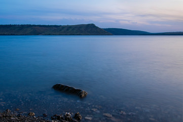 peaceful long exposure nature landscape photography of stone shore line calm river water fuzzy surface and horizon background hill land in twilight time