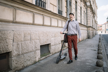 Fototapeta na wymiar Smiling guy walking on sidewalk with scooter. Happy man with moustache wearing elegant clothes walking on sidewalk and holding electric scooter.