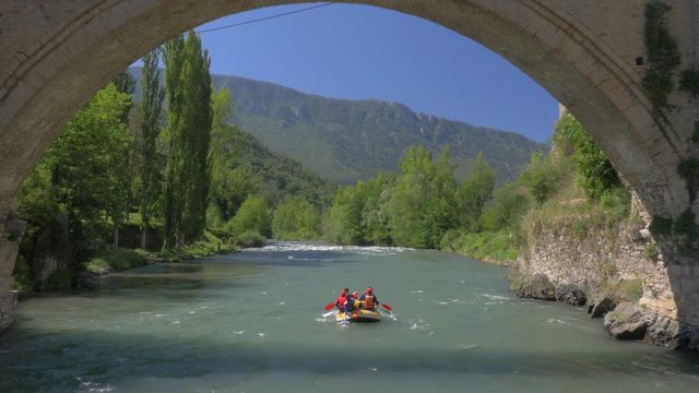 AERIAL DRON SHOT FOLLOWING A RAFTING BOAT WITH PEOPLE ROWING, DESCENDING THROUGH A RIVER AND BELOW A MEDIEVAL BRIDGE