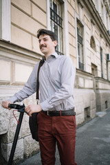 Fototapeta na wymiar Man holding scooter rudder while standing in front of building. Happy man with moustache in elegant outfit standing on sidewalk with scooter.