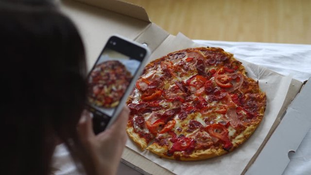Woman taking picture of italian pizza with smartphone lying in bed, pizza served in a cardboard box, fast food delivery.