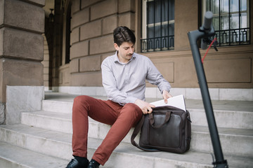Young man pulling laptop from bag while sitting onstairs outdoor. Guy getting computer from his bag while sitting on stairs in front of building.