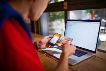 Man online booking via cell telephone and using credit debit card for payment while sitting with pc laptop computer in coffee shop during recreation time