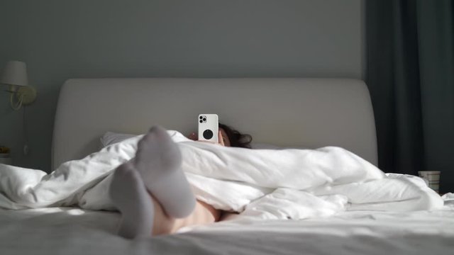 Woman lying in bed and using her smart phone, female feet in white ankle socks under blanket close-up.