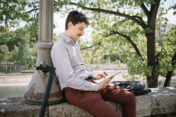 Fototapeta premium Profile of man working on tablet while sitting with scooter outdoor. Young man with moustache holding tablet while sitting on fence and leaning on pillar.