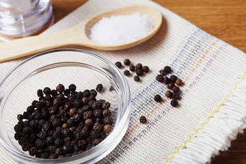 Fototapeta na wymiar black pepper peas in a glass bowl on a linen napkin, salt mill on a wooden kitchen table, soft focus