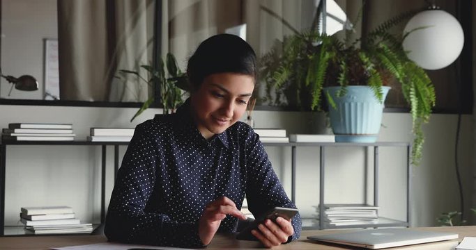 Professional indian businesswoman holding modern smart phone texting message in office. Young female employee using modern mobile technology business app, typing email on cell sitting at work desk.