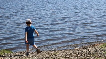 Fototapeta na wymiar a boy in blue clothes from the back walks along the rocky shore of a lake or river to the small waves rolling on the stones