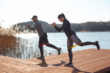 An adult man and woman in sports clothing perform exercises with fitness elastic bands on a pier on the lake. The concept of a healthy lifestyle. Side view