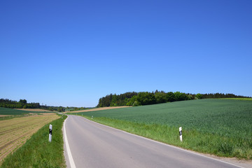 A narrow country road winds through romantic green Bavarian countryside, with blue skies, in spring
