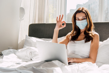 female freelancer in medical mask showing OK sign while working on laptop during self isolation in bed