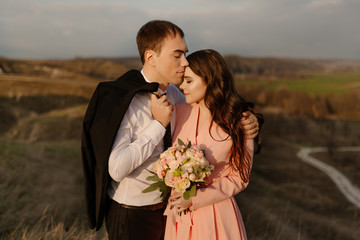 Groom kisses bride on the forehead, against the background of a beautiful view of nature at sunset
