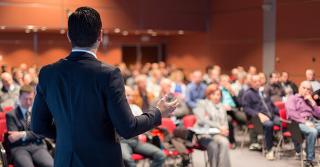 Speaker giving a talk on corporate business conference. Unrecognizable people in audience at...